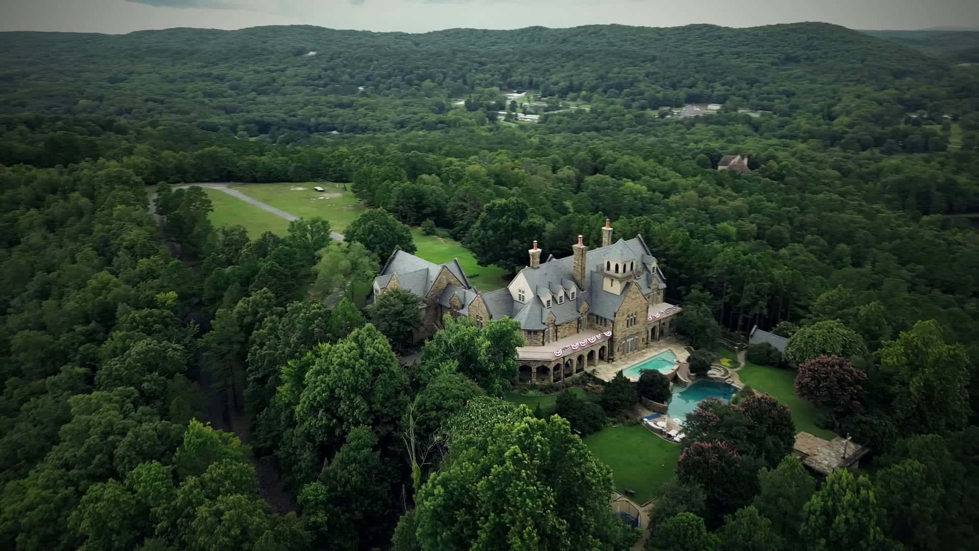 Aerial view over the wooded hills of Central Arkansas toward a stone estate with turrets and a pool